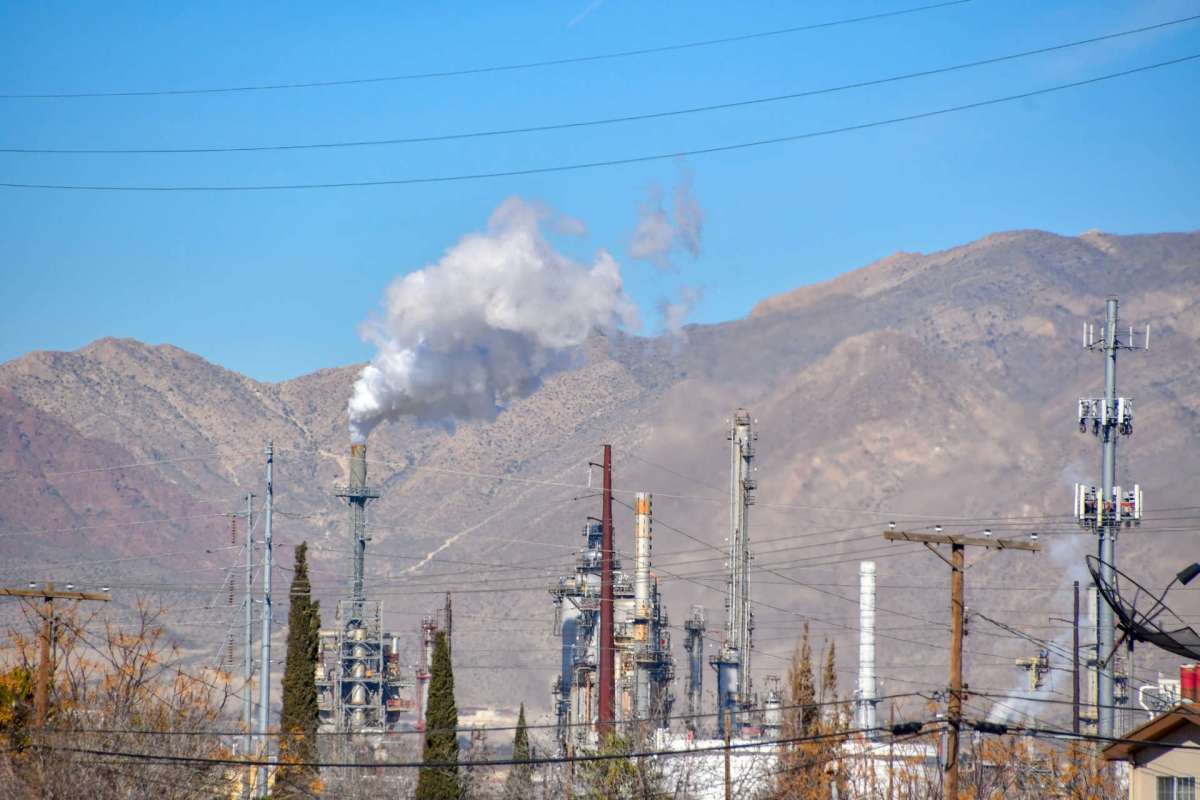 Industrial facilities with smoke stacks and mountains in the background, surrounded by power lines and trees.