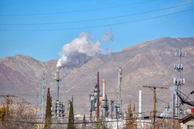 Industrial facilities with smoke stacks and mountains in the background, surrounded by power lines and trees.