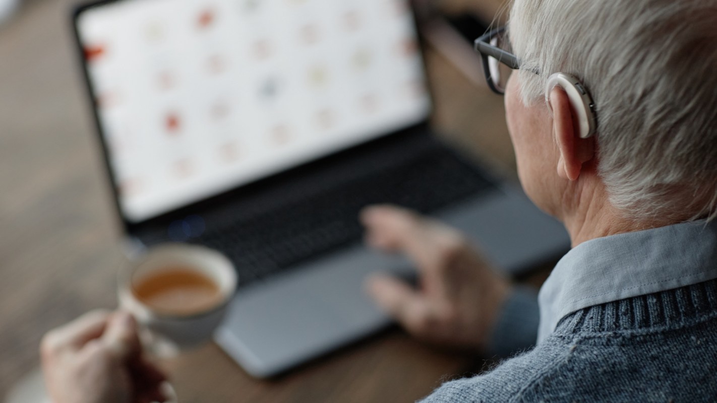 A person with a hearing aid uses a laptop while holding a cup of tea.