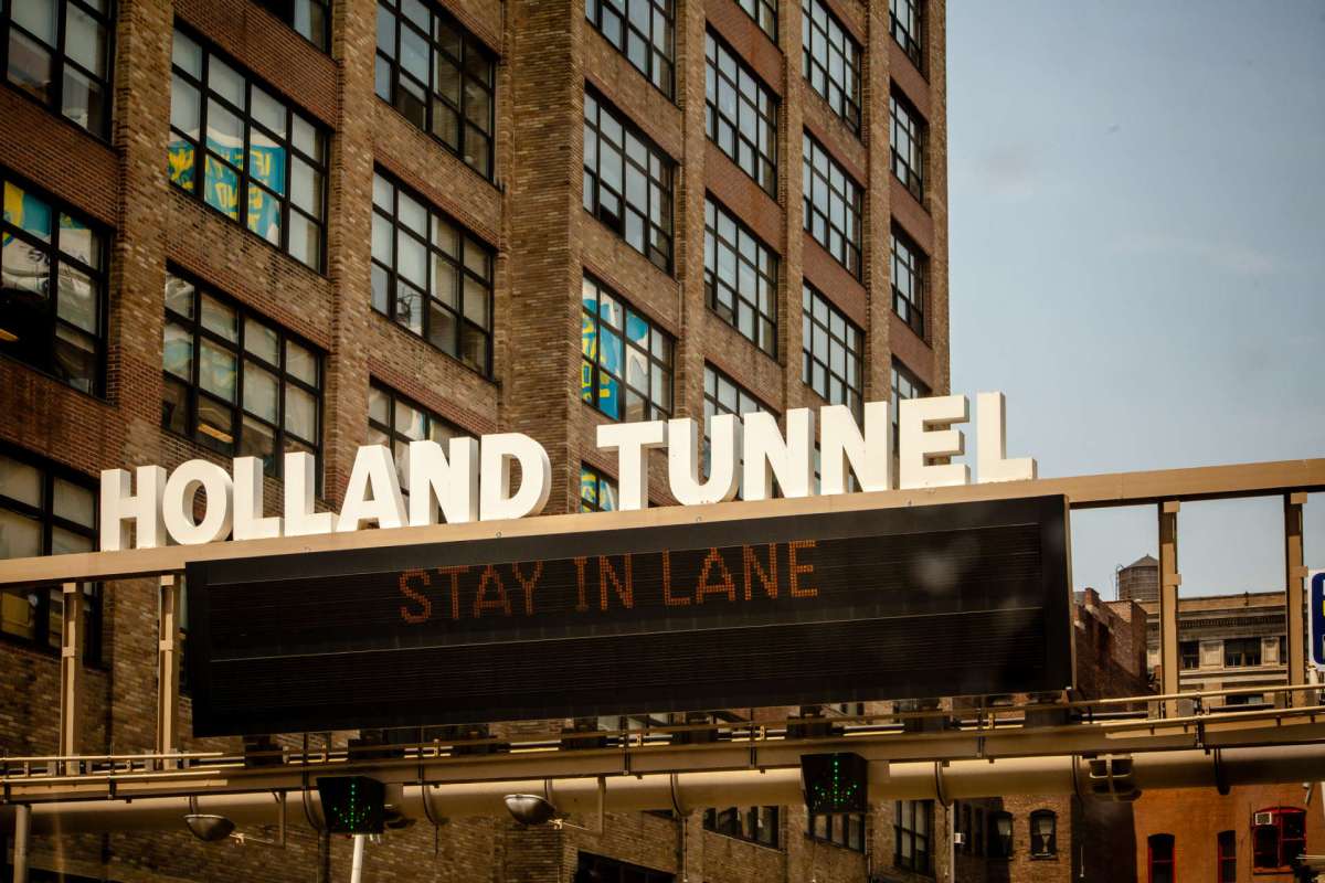 Sign for the Holland Tunnel with a message to stay in lane, next to a brick building with windows.