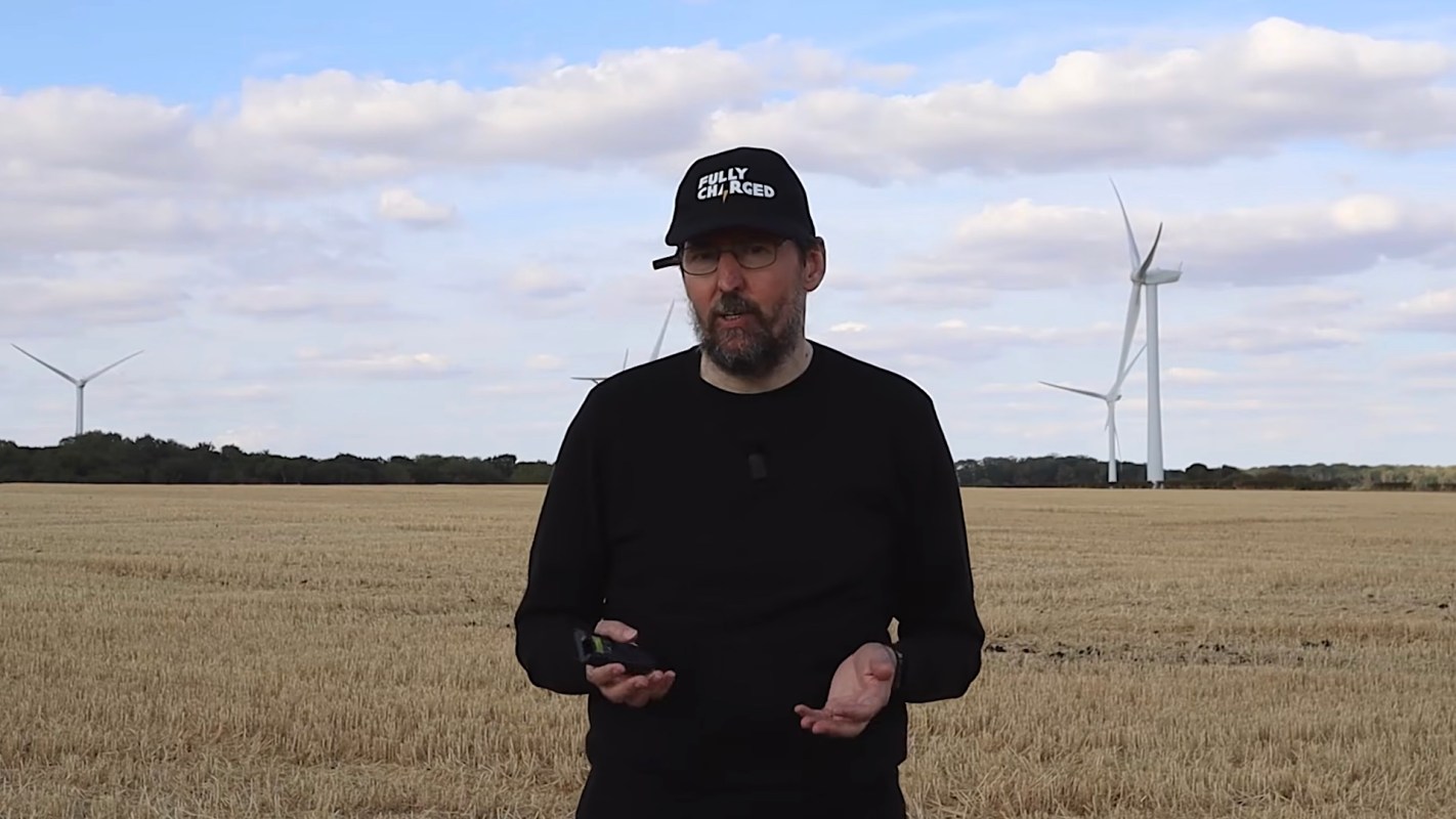A man stands in a field with wind turbines in the background under a cloudy sky.