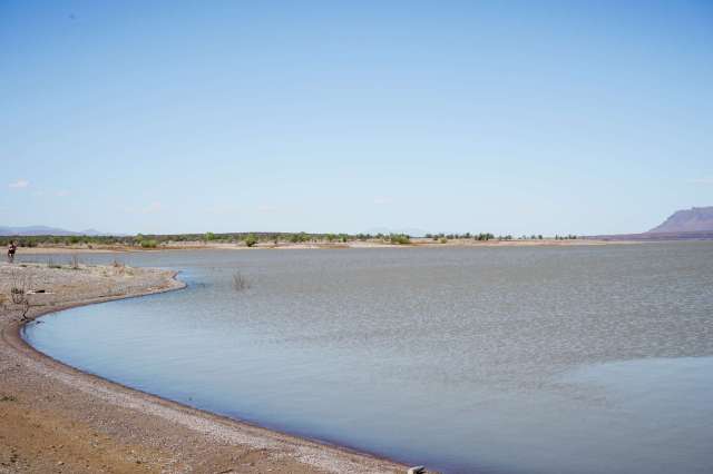 A tranquil lake surrounded by a sandy shore and distant mountains under a clear blue sky.