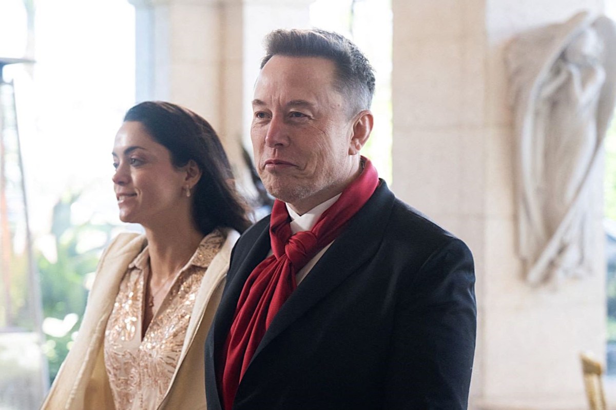 Elon Musk wearing a black suit with a red scarf and a woman walk through a walkway