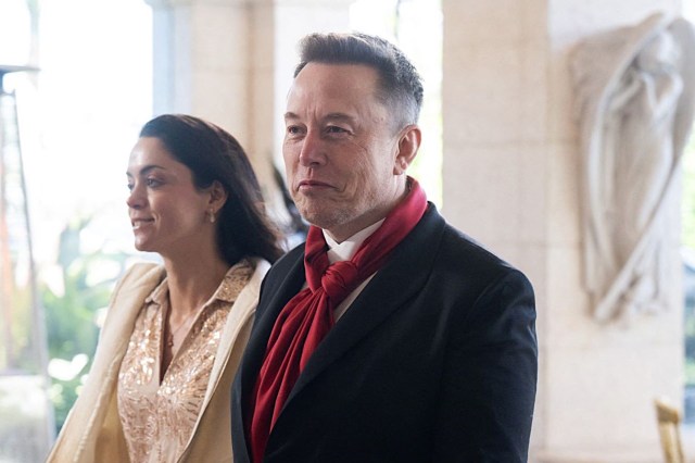 Elon Musk wearing a black suit with a red scarf and a woman walk through a walkway