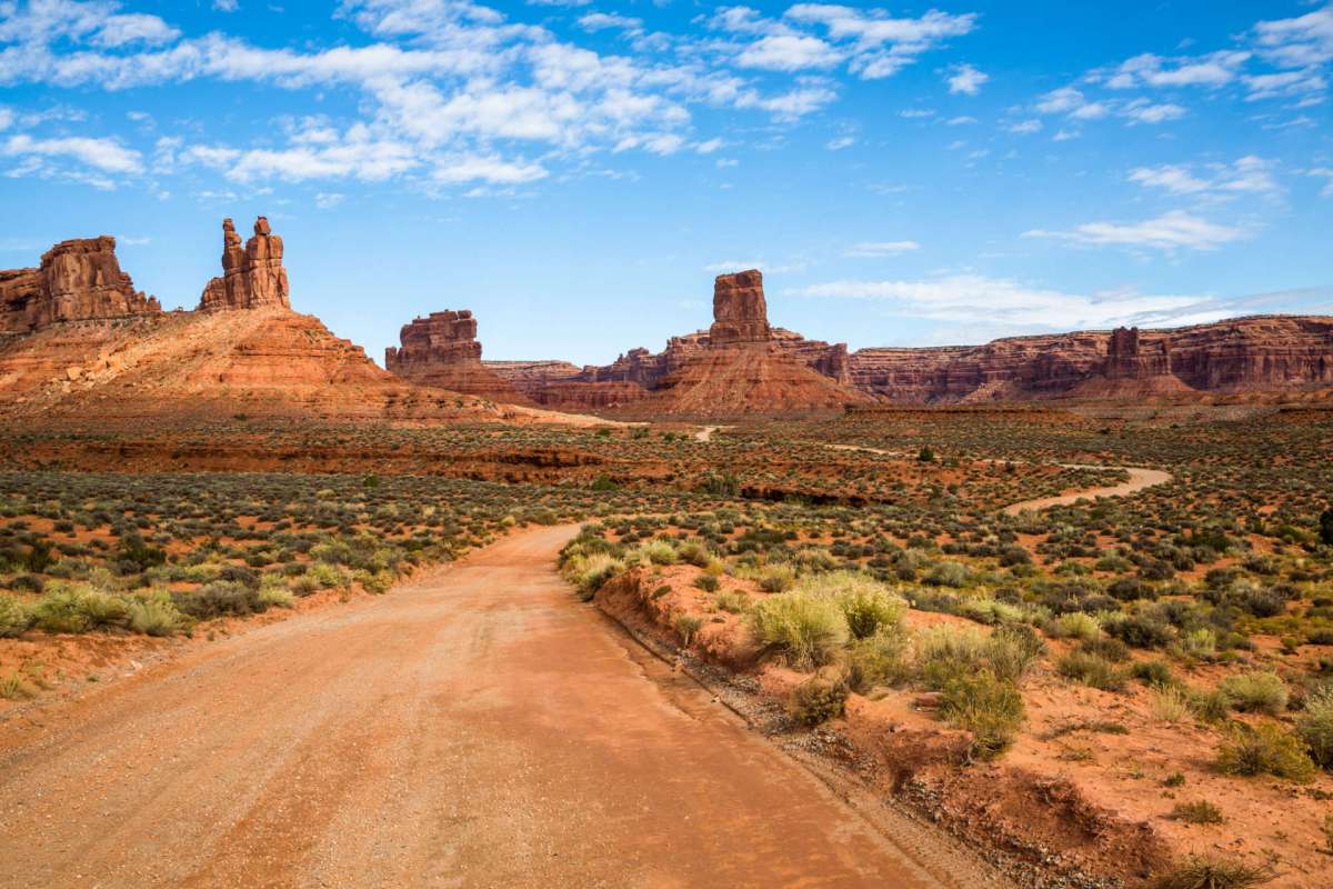 A dirt road winds through a desert landscape with red rock formations and sparse vegetation under a blue sky.