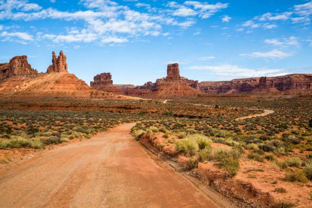 A dirt road winds through a desert landscape with red rock formations and sparse vegetation under a blue sky.