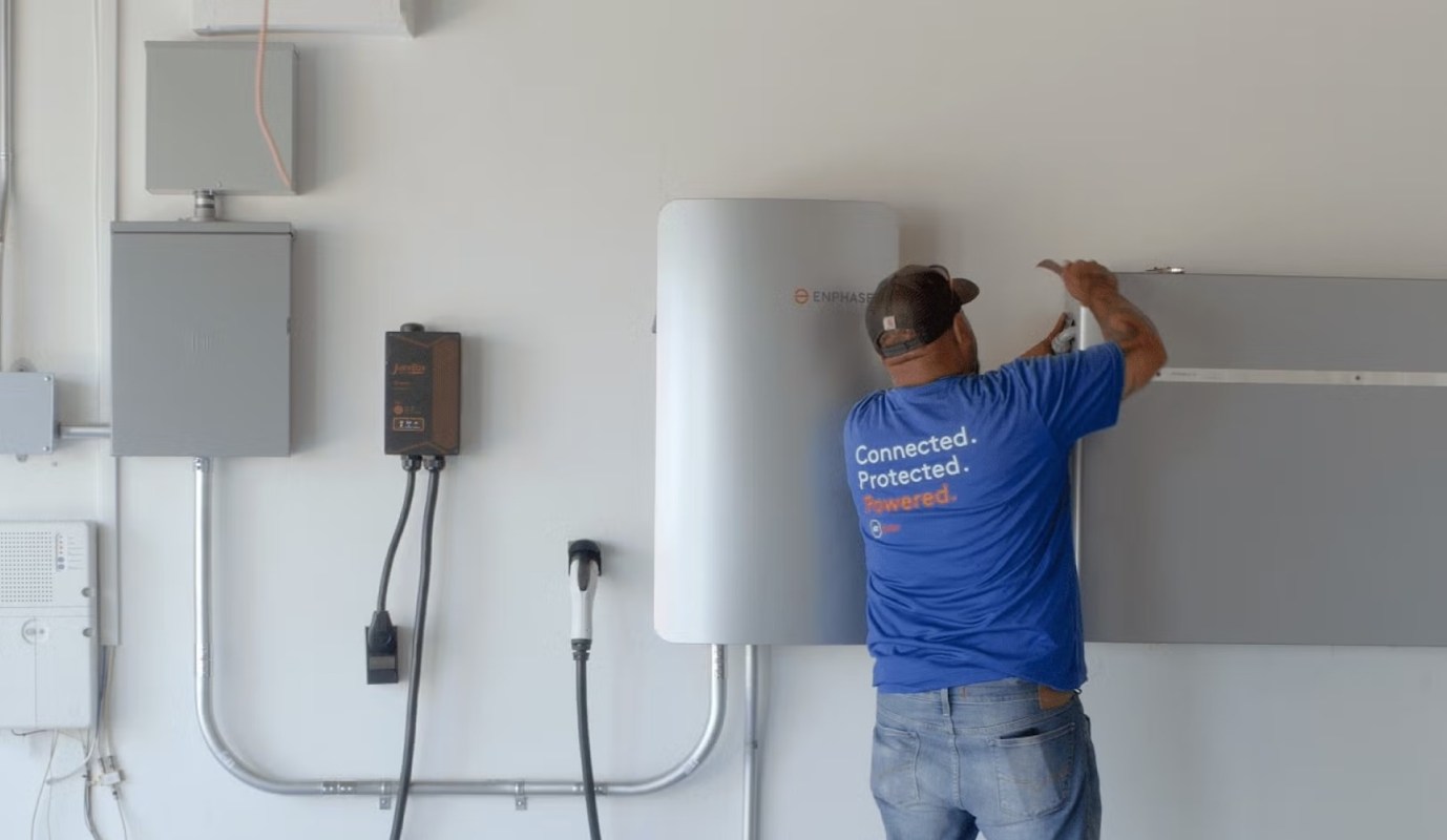 A person installs Enphase equipment on a wall in a space with electrical panels and a charging station.