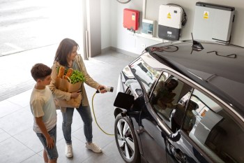 A woman plugs in an electric vehicle while a boy stands beside her holding a bag of groceries.
