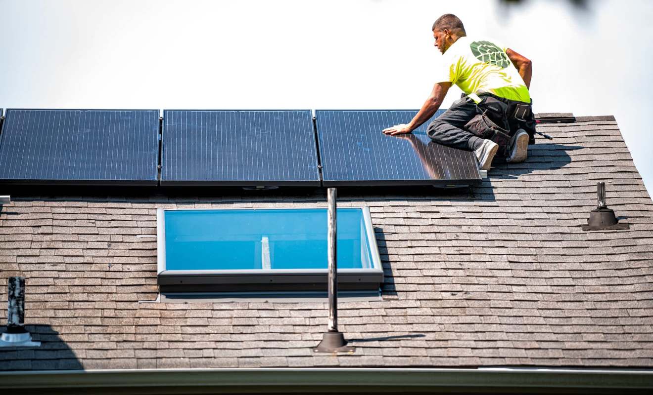 A worker installs solar panels on a sloped roof with a skylight.