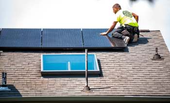 A worker installs solar panels on a sloped roof with a skylight.