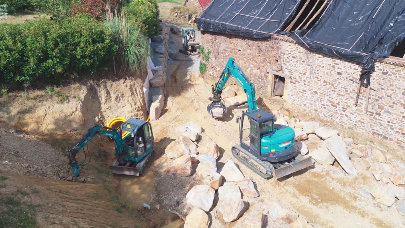 Two excavators work on a construction site with large rocks and a partially covered building in the background.