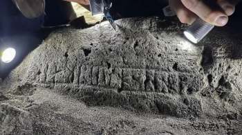 An archaeologist examines engraved markings on a rocky surface using a flashlight.