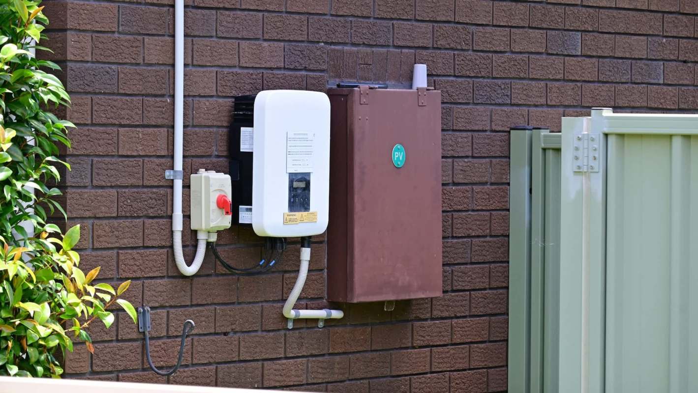 A solar inverter and electrical box mounted on a brick wall next to a green garden fence.