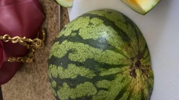 A watermelon sits on a cutting board next to a handbag.