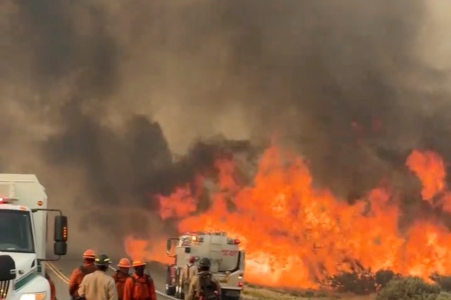 Firefighters and fire trucks are present near a large wildfire with thick smoke and flames rising in the background.