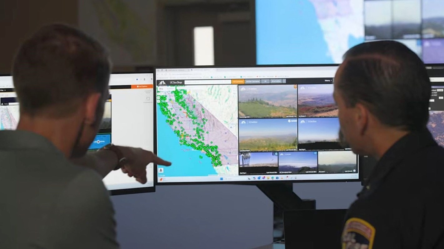 Two individuals examine multiple computer screens displaying maps and live video feeds.