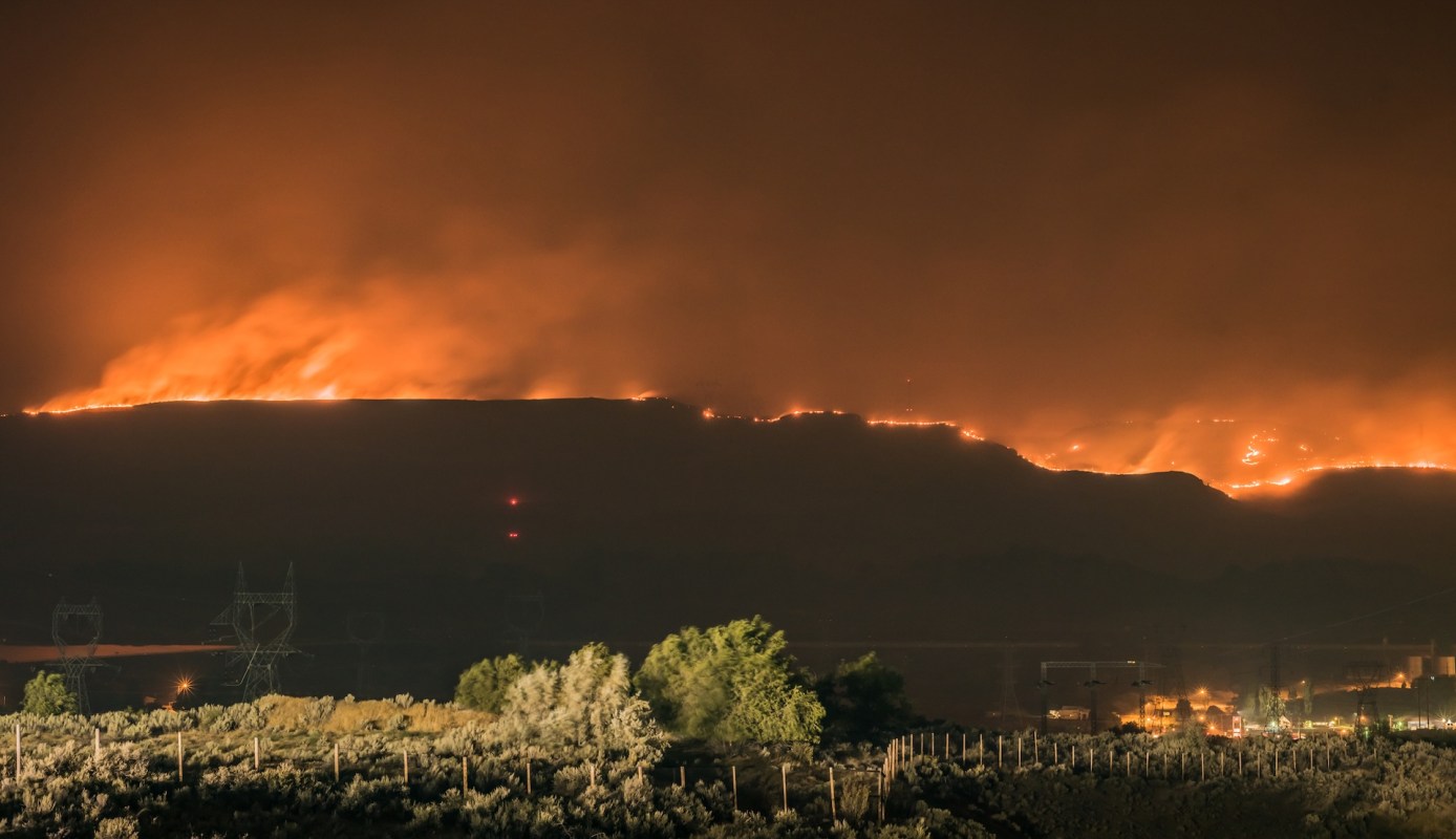 A hillside is engulfed in flames under a smoky, orange sky.