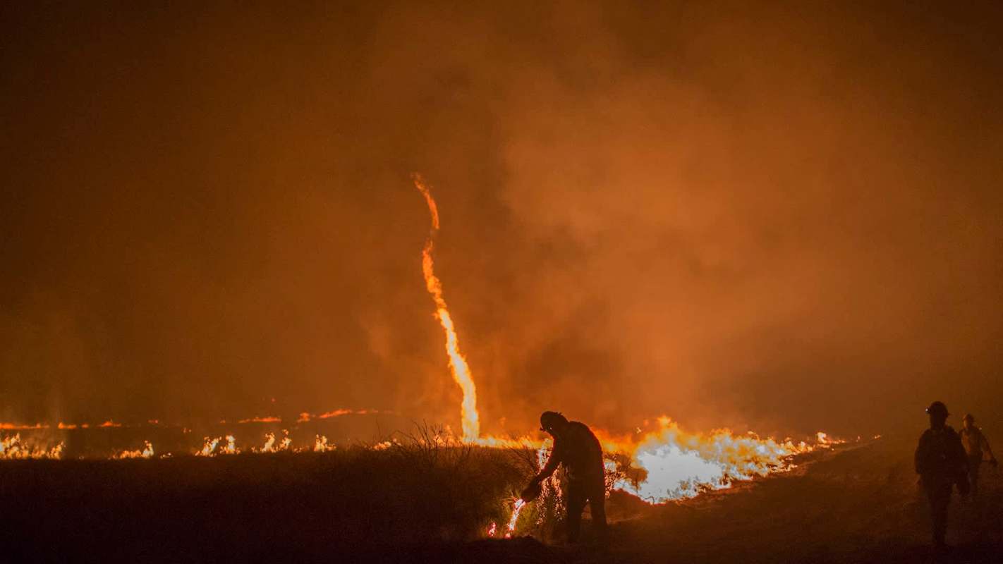 Firefighters manage a wildfire, including flames in the shape of a tornado funnel, and smoke in a grassy area.