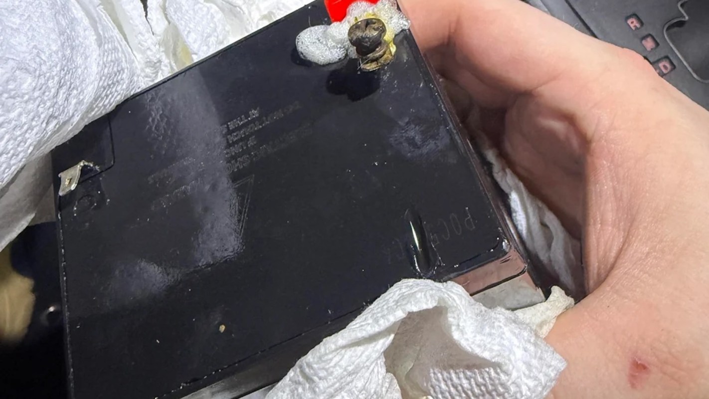 A hand holds a black battery with foam around a terminal, framed by paper towels.