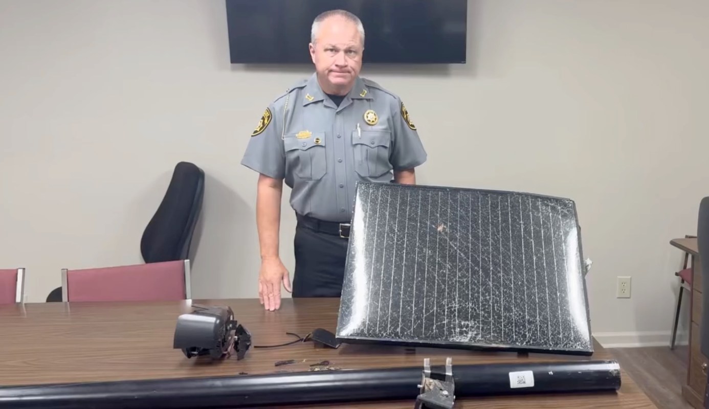 A police officer stands behind a table with a vandalized Flock camera.