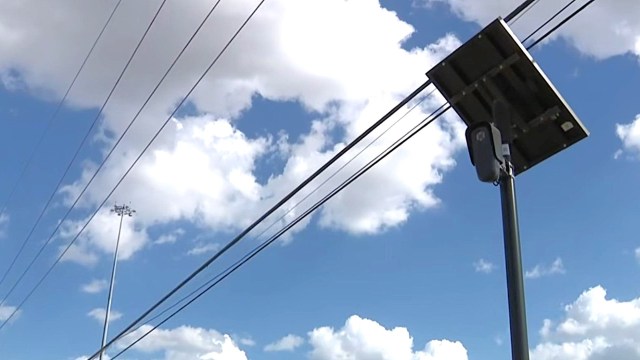 A camera with solar panel mounted on a pole beneath a blue sky with clouds and power lines.