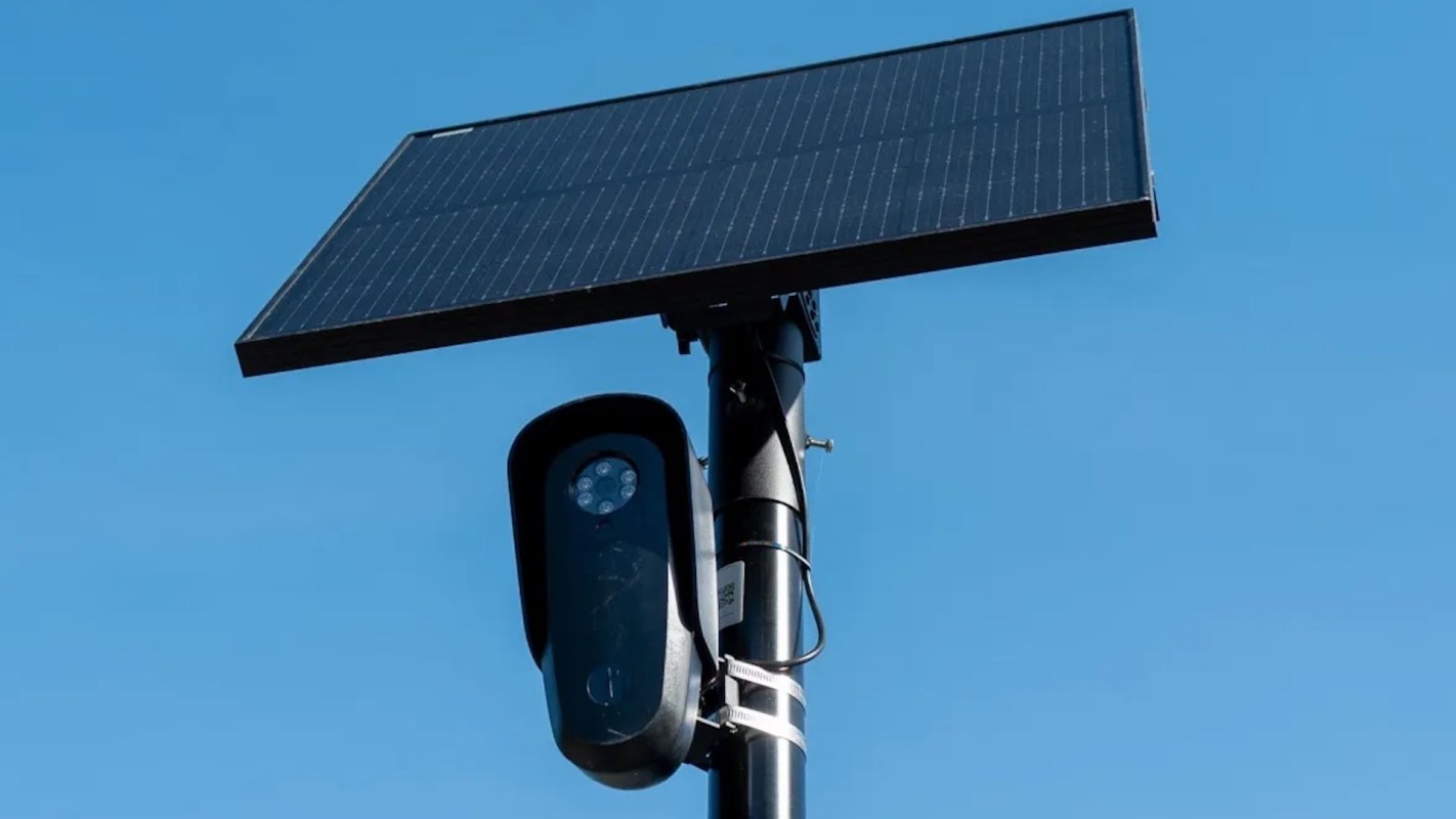A solar panel mounted on a pole with a camera attached below it against a blue sky.