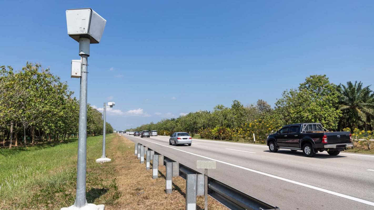 Traffic cameras are mounted along a highway lined with trees and vehicles traveling in both directions.