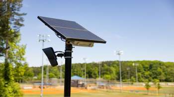 A solar panel and light sensor mounted on a pole overlooking a sports field.