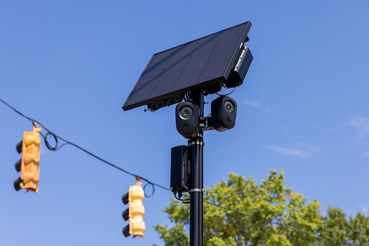 A solar-powered surveillance camera system mounted on a pole with traffic lights in the background.