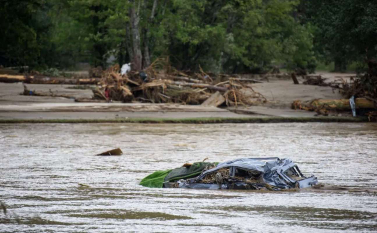 A submerged car in a river with debris and trees in the background.