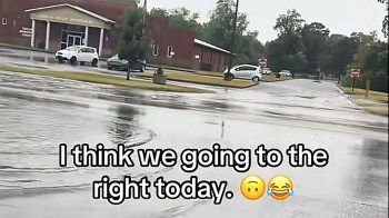 A wet street near a building with a sign and several parked cars on a rainy day.