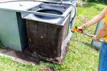 A person uses a hose to wash the exterior of an air conditioning unit on a grassy area.