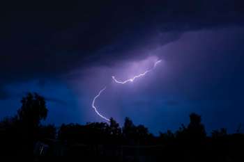 Lightning strikes through dark, stormy clouds illuminated by a blue sky.