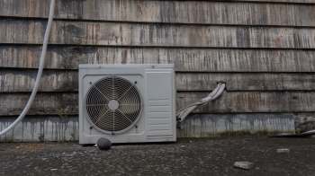 An air conditioning unit against a weathered wooden wall with water stains and a few stones on the ground.