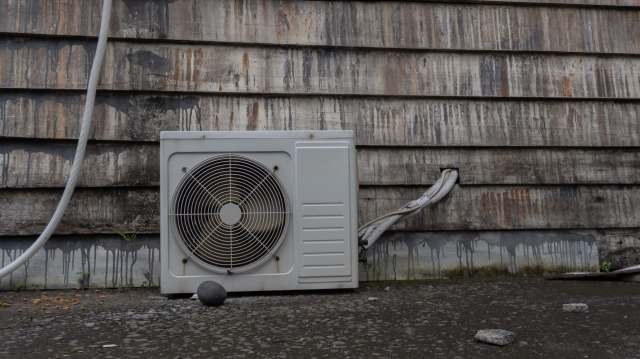An air conditioning unit against a weathered wooden wall with water stains and a few stones on the ground.
