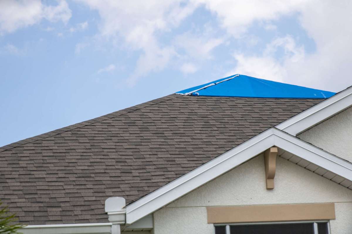 A roof with a blue tarp covering part of the shingles under a cloudy sky.