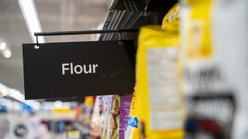 A sign labeled "Flour" hangs above bags of flour on a grocery store shelf.