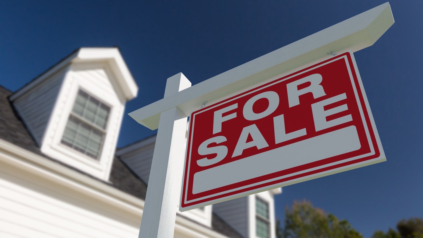 A "For Sale" sign in front of a house against a clear blue sky.