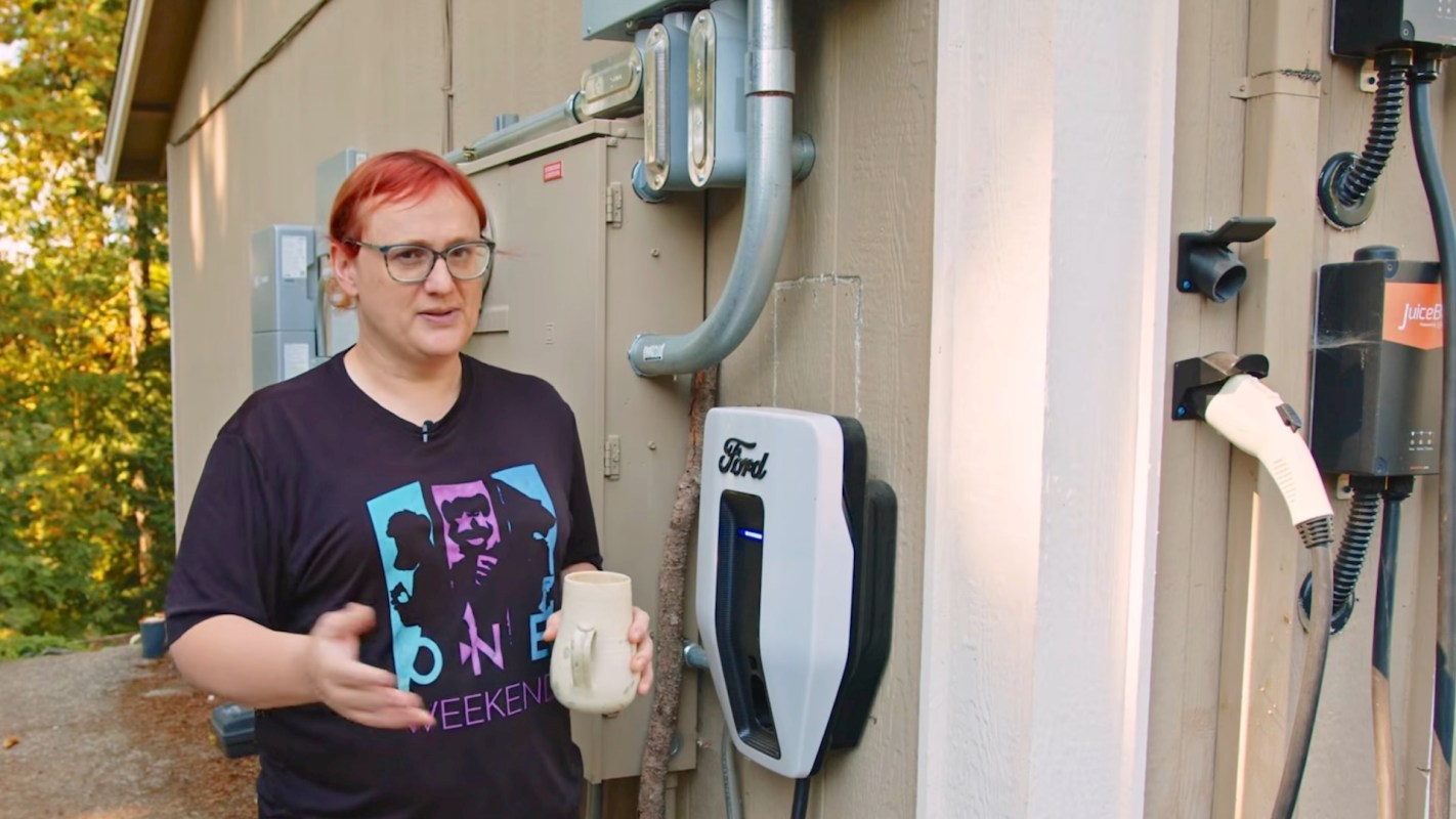 A person stands next to an electric vehicle charger attached to a wall, holding a ceramic mug.