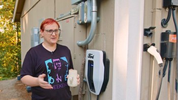 A person stands next to an electric vehicle charger attached to a wall, holding a ceramic mug.