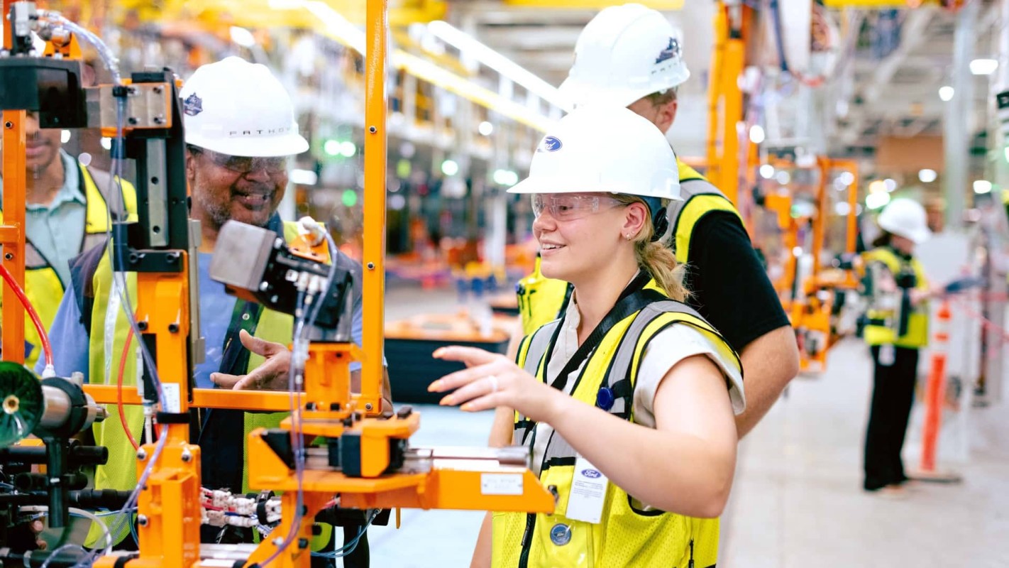 A group of workers in hard hats and safety vests observes machinery in a factory setting.