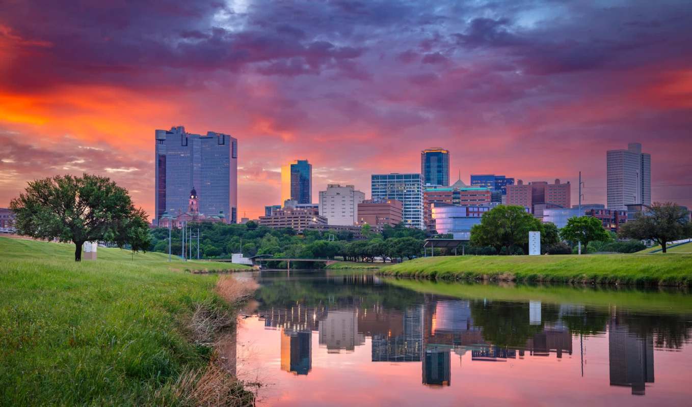 A city skyline at sunset reflected in a calm river with green grass and trees in the foreground.