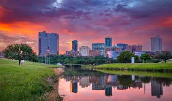 A city skyline at sunset reflected in a calm river with green grass and trees in the foreground.