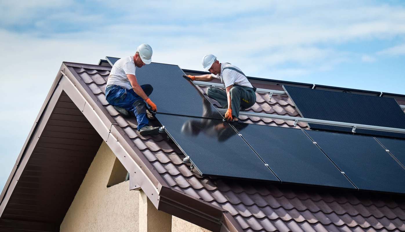 Two workers install solar panels on a brown-tiled roof under a blue sky.