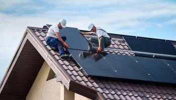 Two workers install solar panels on a brown-tiled roof under a blue sky.