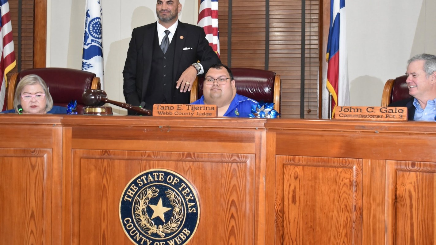 Four officials sit at a wooden dais with flags in the background, a gavel and nameplates are visible.