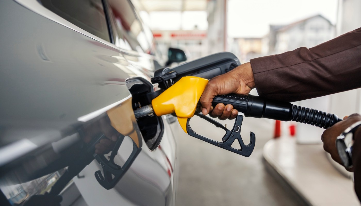 A hand holds a fuel pump nozzle while refueling a vehicle at a gas station.