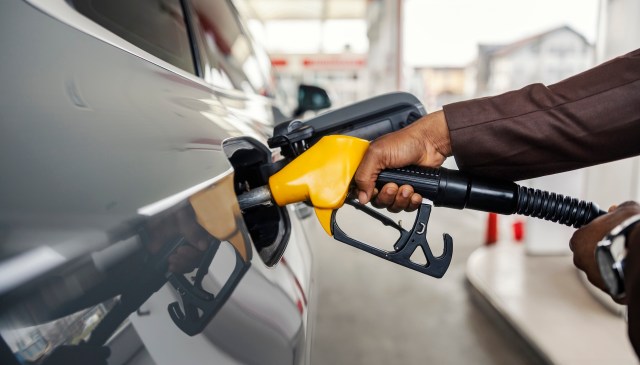 A hand holds a fuel pump nozzle while refueling a vehicle at a gas station.