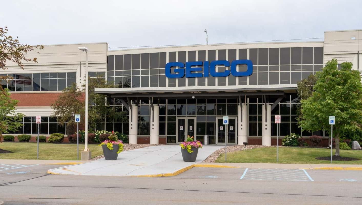 A GEICO office building with glass doors and flower planters in front.