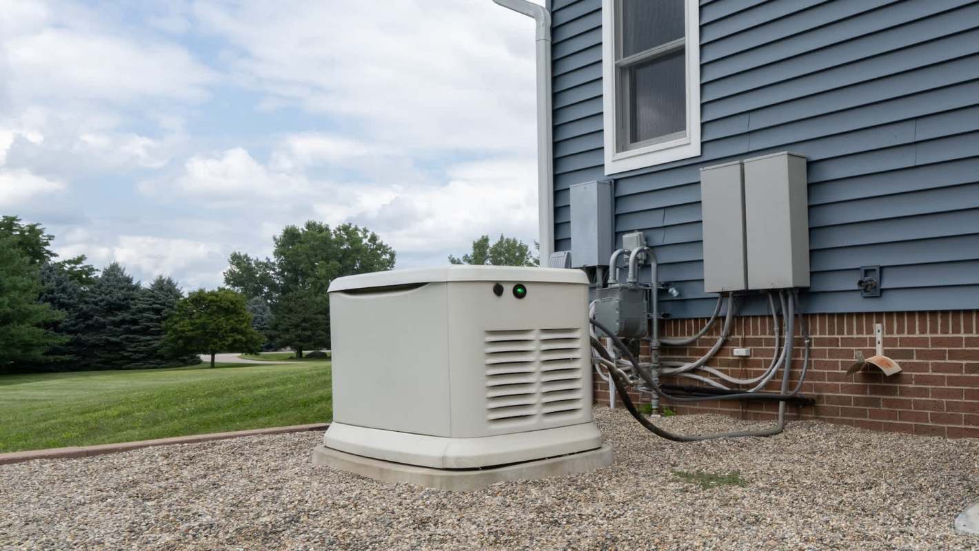 A generator is located beside a house, connected to electrical equipment and surrounded by gravel.